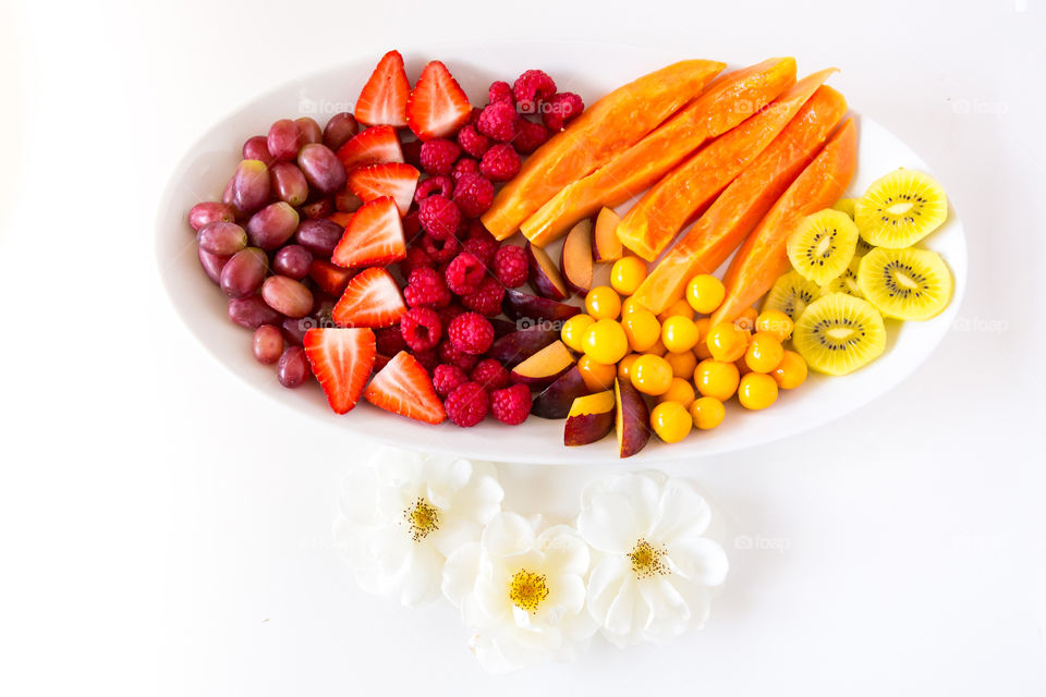 Sliced summer fresh fruit platter on white plate with variety of red orange and yellow fruits. White flowers with a white background. Fruits include strawberries gooseberries grapes raspberries papino and kiwi. Fresh healthy food.