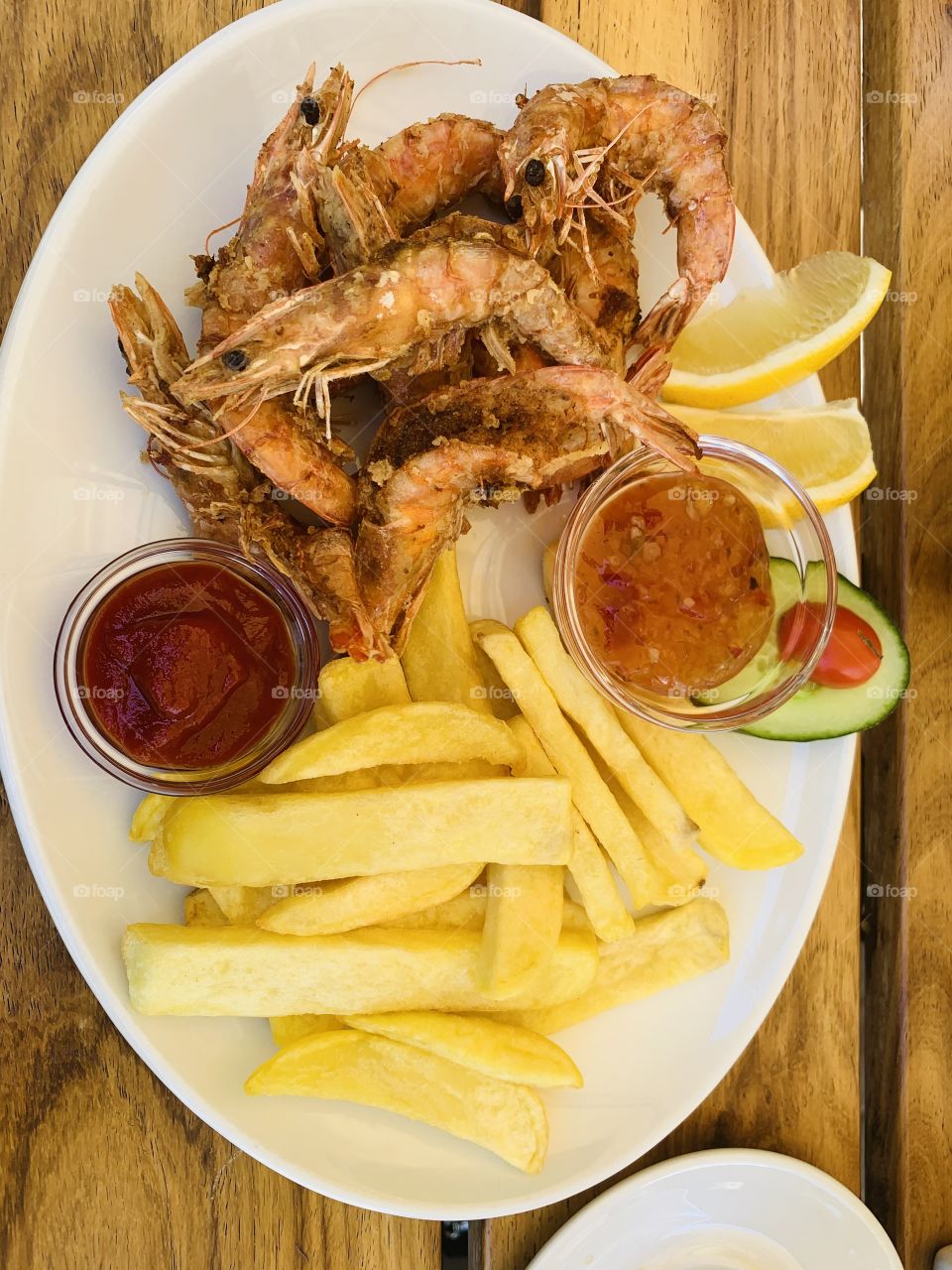 A white lunch dish of deep fried prawns, lemon slices, French fries, sweet chili sauce and catchup. 