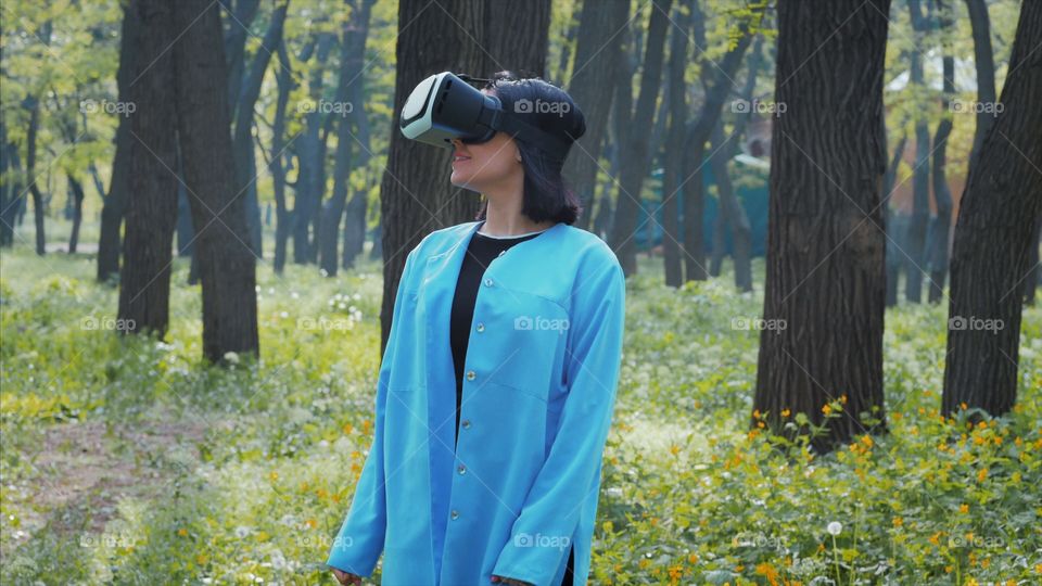 Portrait of trendy woman in blue coat using virtual reality glasses simulator in the park or forest at spring 
