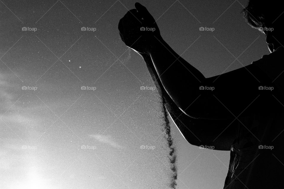 Silhouette of person letting sand funnel out of their hands with a few wispy clouds in the sky in the background