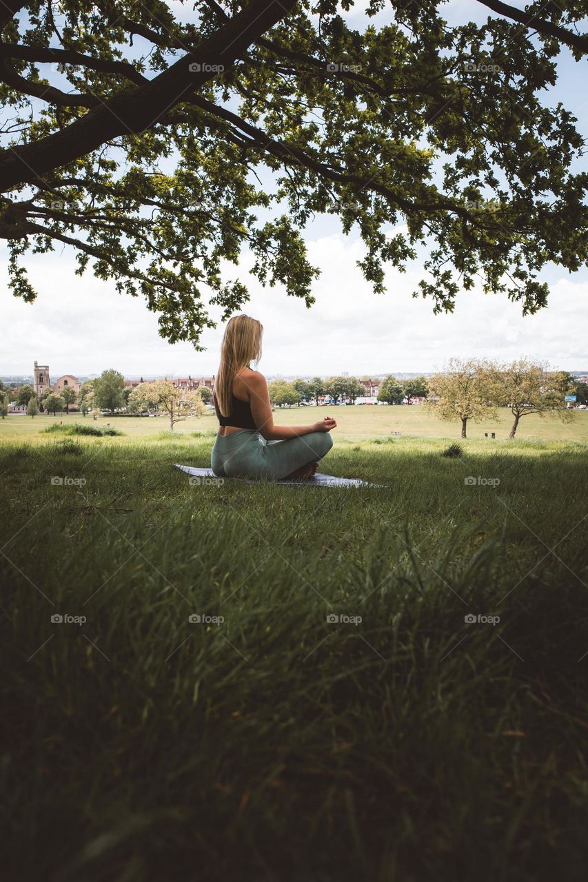 Woman Meditates Cross Legged Under A Tree Photo
Woman is sitting under a tree on a yoga mat facing away from the camera. She has her legs crossed and her hands on her knees.