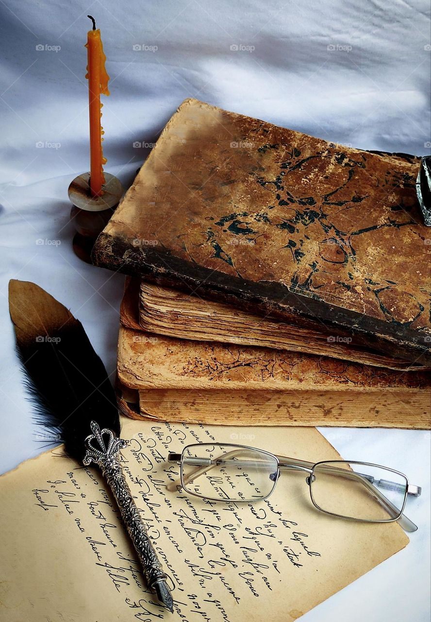 A sleepless night. Letter. In the background are brown antique books and an extinguished candle stands nearby. In the foreground lies a sheet of paper with handwritten black text, on which lie glasses and an ink pen. The background is light