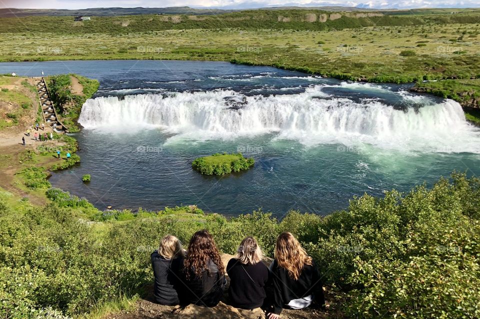 Four friends sit in the green grass overlooking a wide, beautiful waterfall. The water is blue and the sun is out!