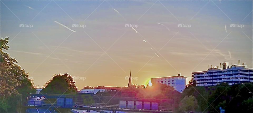 A vivacious “Munich” evening with a large orange glowing sun setting gradually behind the horizon in the far distance illuminating all city buildings including the steeples of churches with its warm reddish golden glow. 2022. Hypnotic Productions
