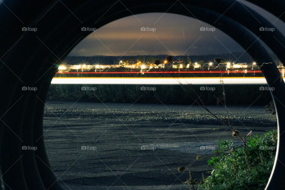 Nighttime shot of a car travelling down a road framed through a circular bicycle rack . Soaking in as much light as possible with wide aperture & long shutter speed and a tripod. Marina & car front & side lights are carried throughout the shot. 