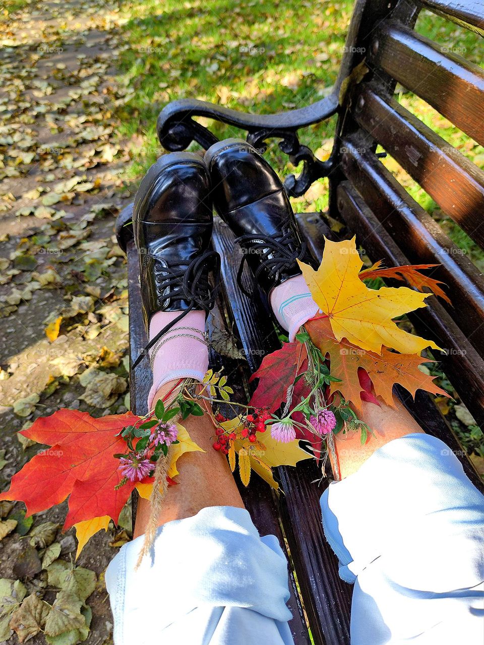 Autumn.  A wooden bench stands among autumn trees.  On the bench are legs in blue trousers, pink socks and black shoes.  Bouquets of multi-colored maple leaves, pink clover and red rowan stick out of the socks.