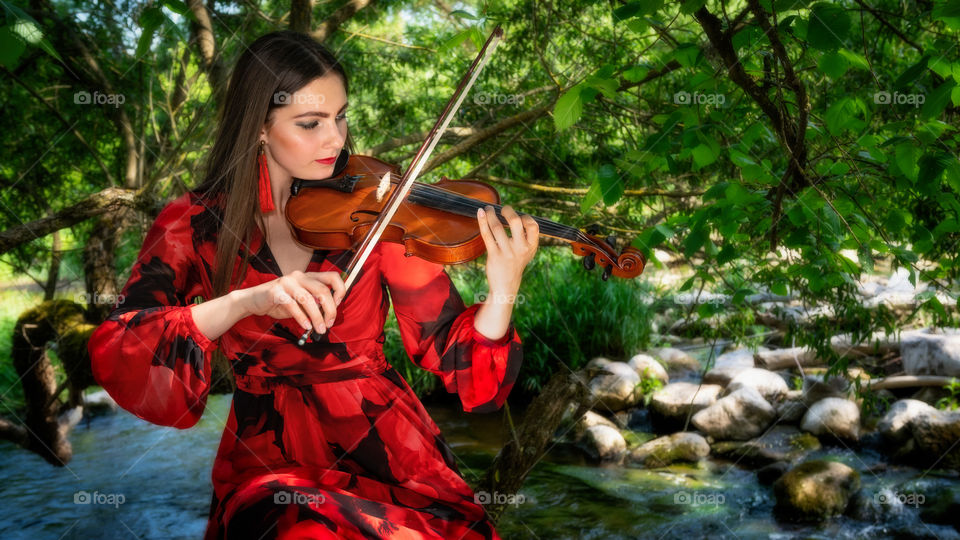 Young, beautiful woman in a red dress playing violin on the riverbank.