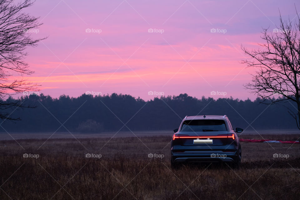 Car stay in the field with a beautiful purple sunrise and trees silhouette on background 