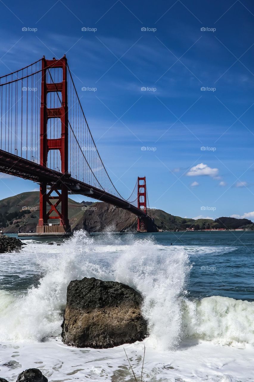 Golden Gate Bridge viewed from Fort Point in San Francisco California with the waves crashing onto the rocks and the clear blue skies above 