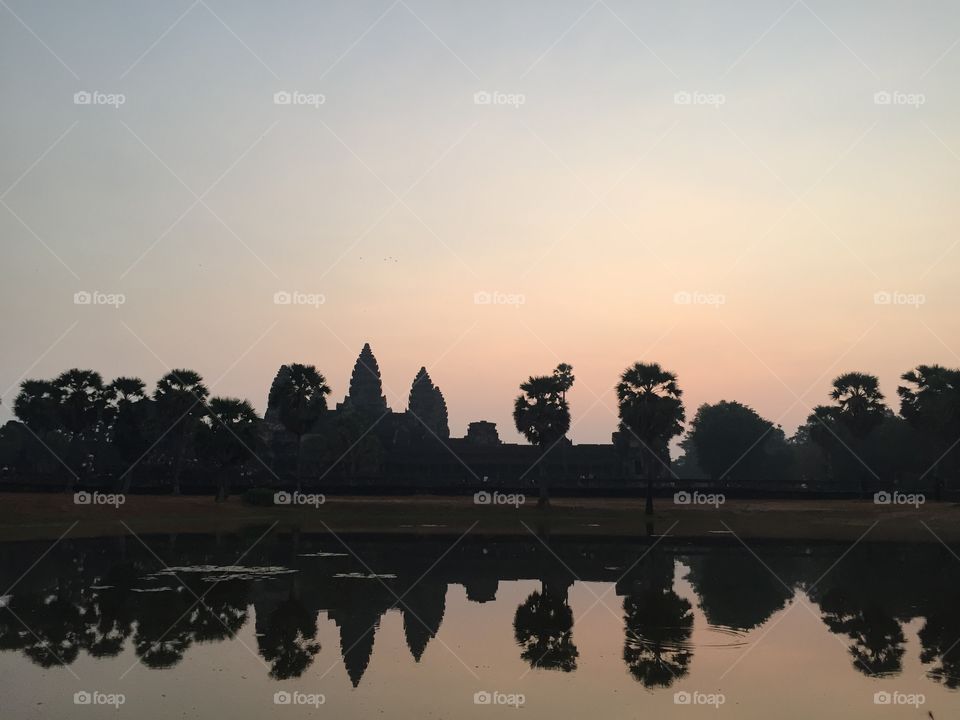 Angkor Wat temple in Cambodia with sunrise. Peaks of temple visible through trees. Reflection of water with lotus pads show the temple. Silhouette of temple shown.