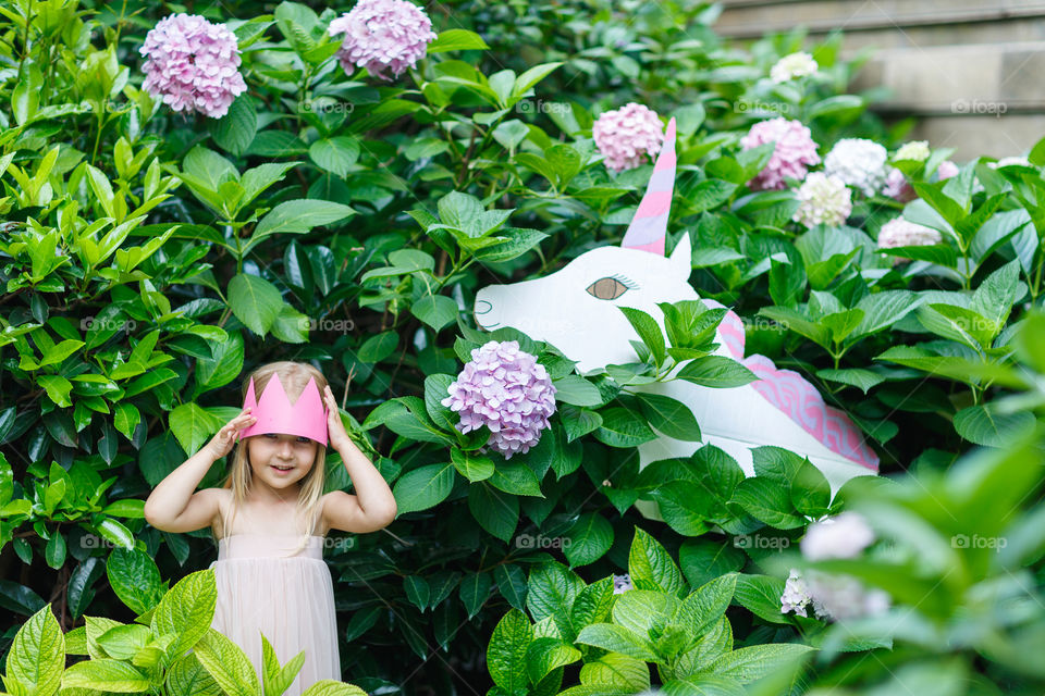 Happy little Caucasian girl with blonde hair and crown standing near unicorn in blooming garden at summer day