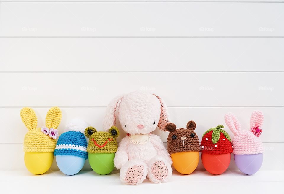 Cute plush bunny doll with colorful Easter eggs with crochet Easter egg cups top in various shapes of bunnies, frog, teddy bear, strawberry and pom pom winter hat on white wooden board.