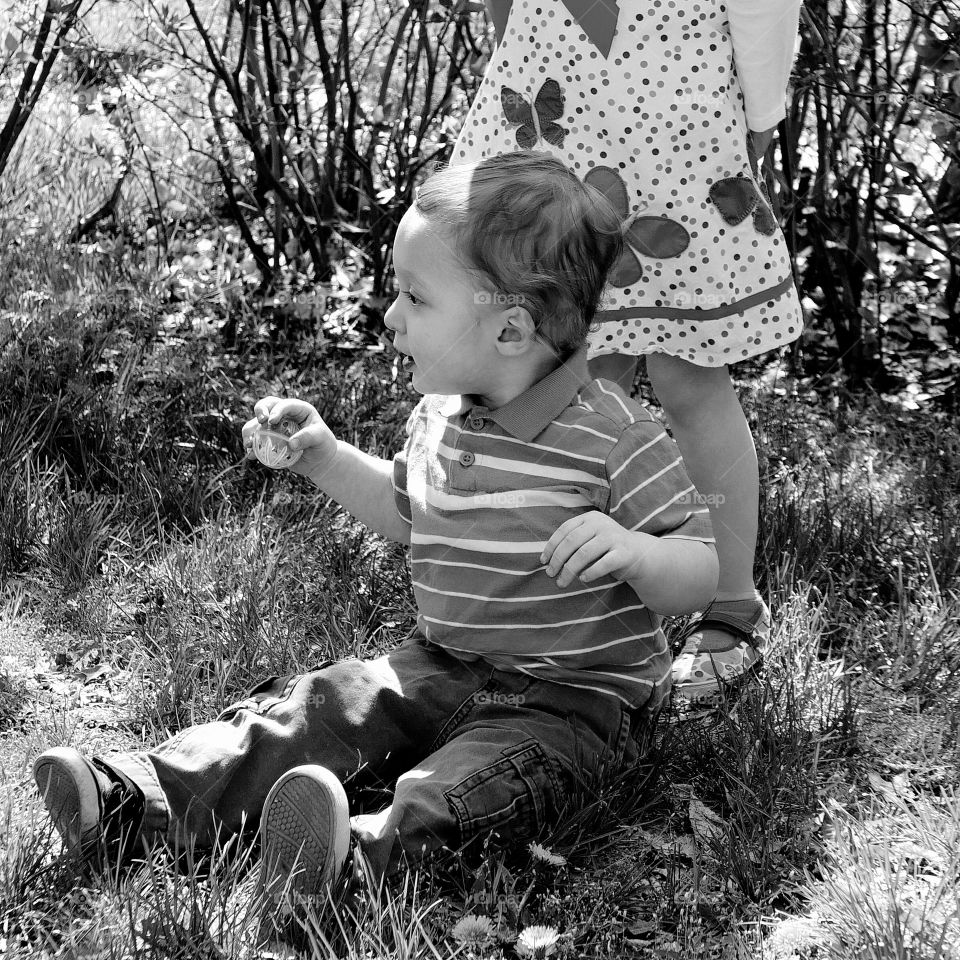 A toddler boy sits at the feet of his sister in the grass during an Easter egg hunt on a sunny spring day. 