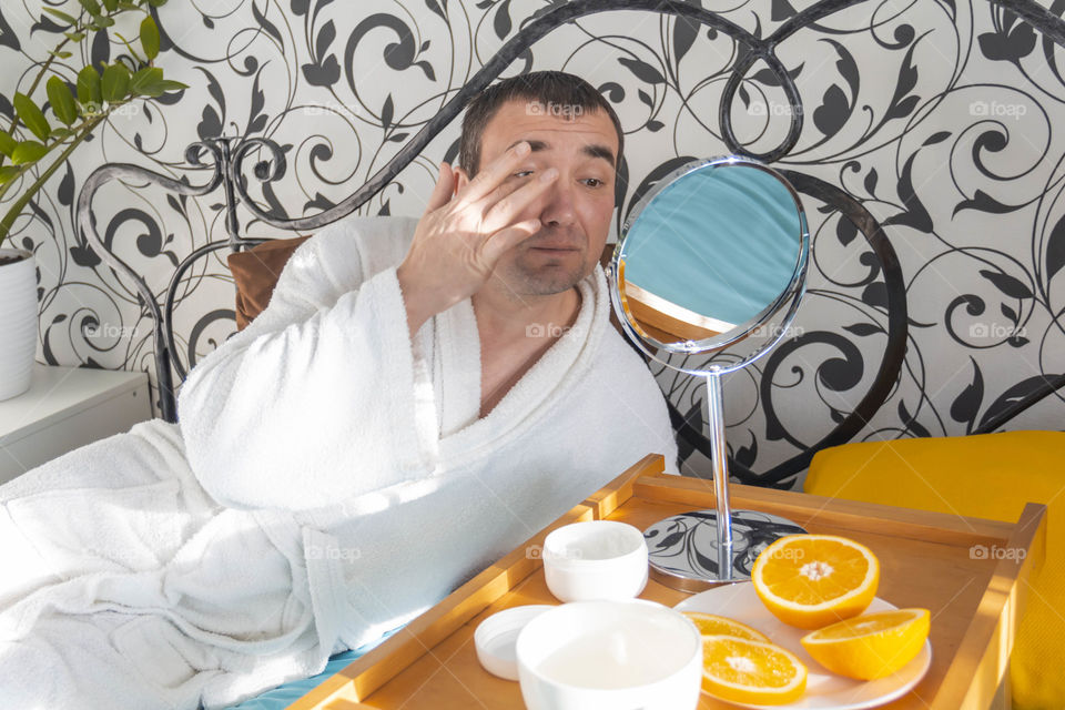 A man and home spa care at home takes care of himself and smears his face with cream, does a manicure at home on a blue background and with oranges and rests.