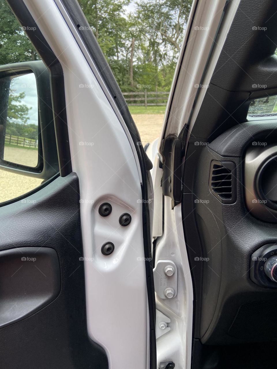 A view of a park through the open door, window and mirror of my Jeep Wrangler. The ground is sandy and trees and a fence are in the background.