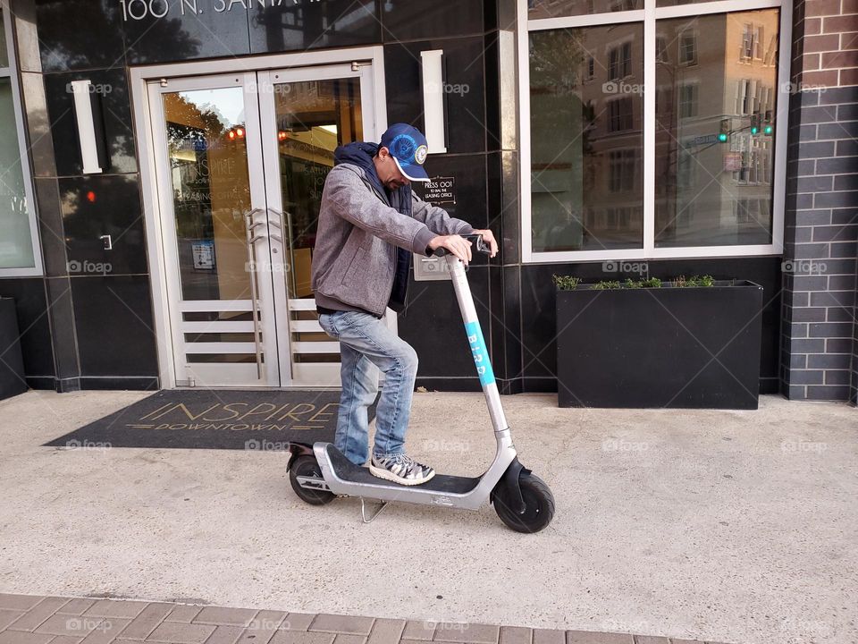 Man checking out a street rental scooter in front of a downtown business public sidewalk .