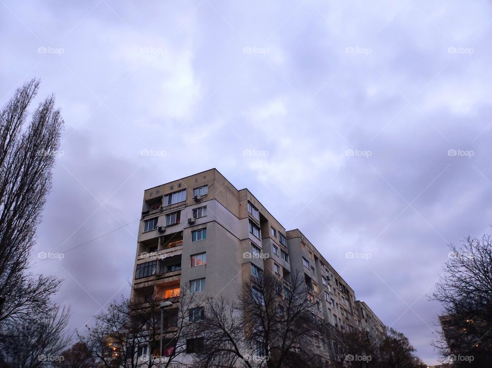 An old Bulgarian apartment building with a cloudy sky overhead and trees around the block