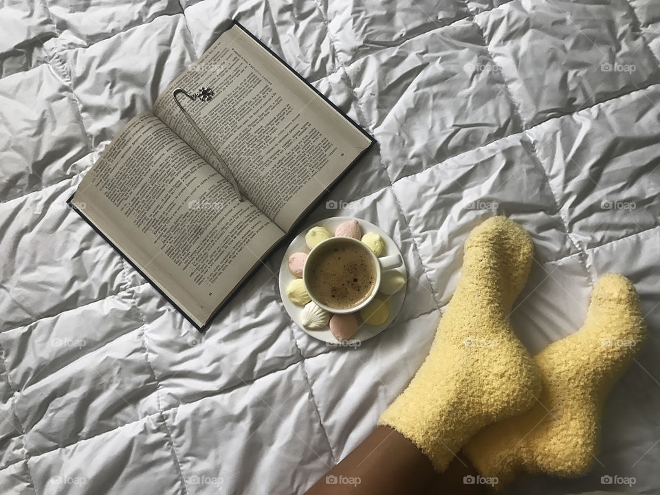 Woman in yellow cozy socks with a cup of coffee reading a book in bed 