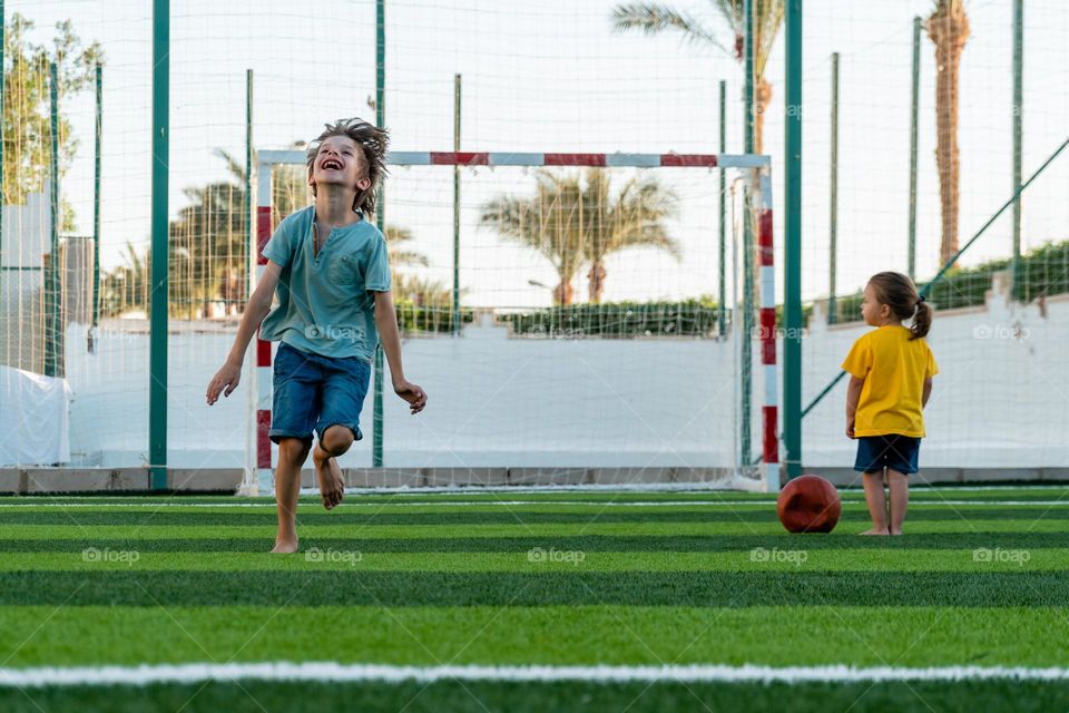 Happy boy running along green lawn and laughing during playing football and little girl standing not far