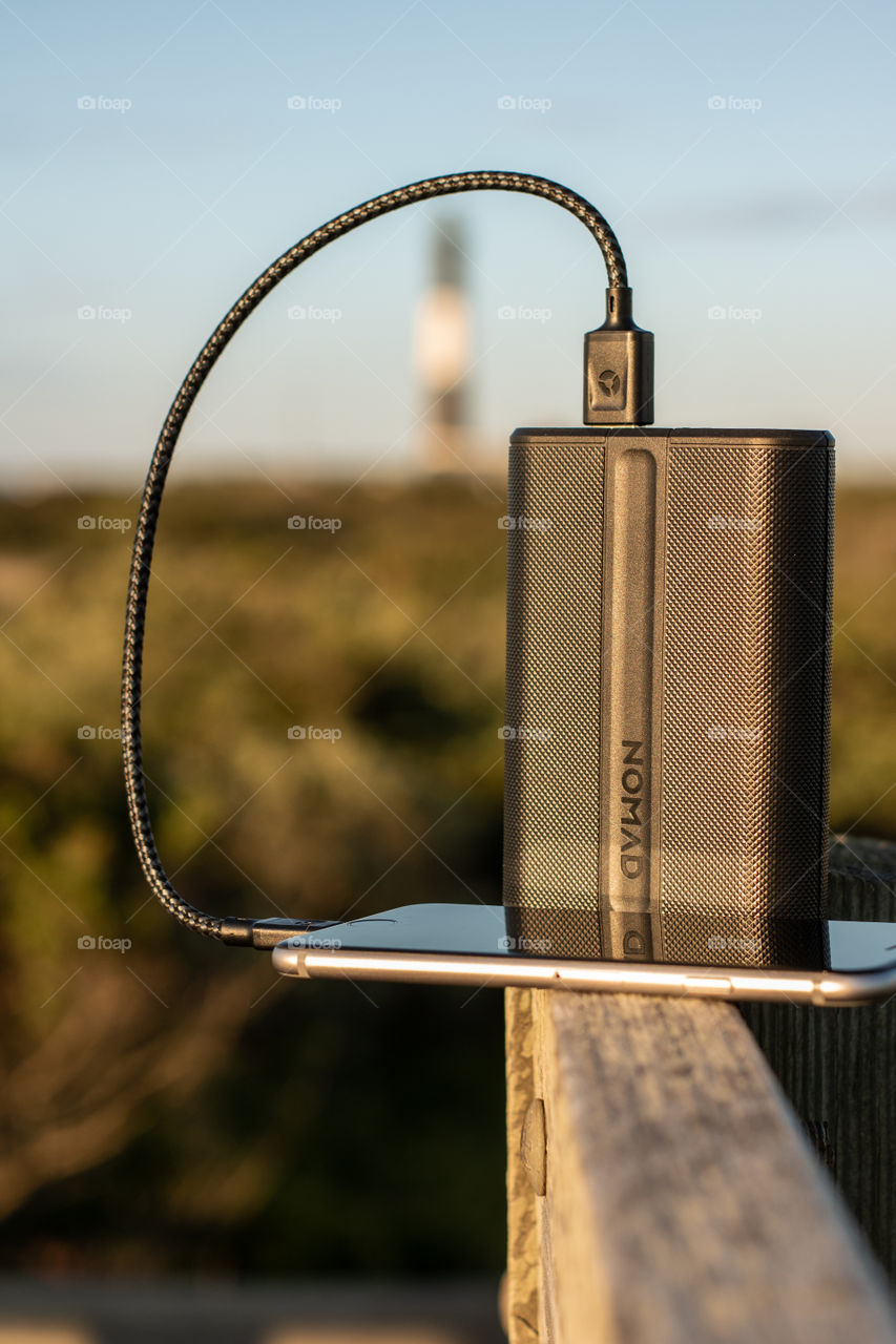 An external battery power bank charging an iPhone - outdoor summer scene on a boardwalk with golden light hitting a lighthouse in the background.