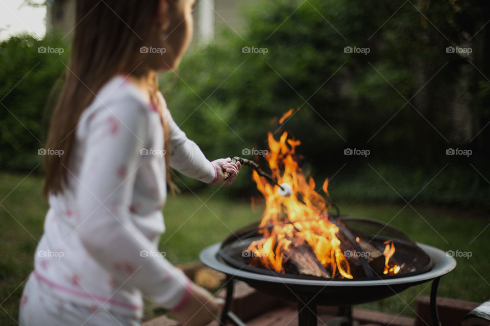 Cute little girl in pajamas roasting a marshmallow on a stick over a fire in the backyard during summer. 