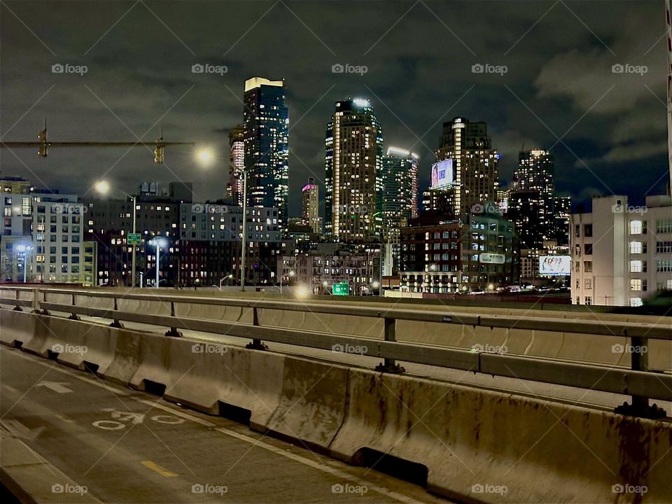 This is the bicycle lane and the car lanes of the “Pulaski Bridge” at “Newtown Creek” in LIC, Queens with a backdrop of the LIC skyline at night. Cars with their bright headlights are zooming past incessantly. 2023. Hypnotic Productions