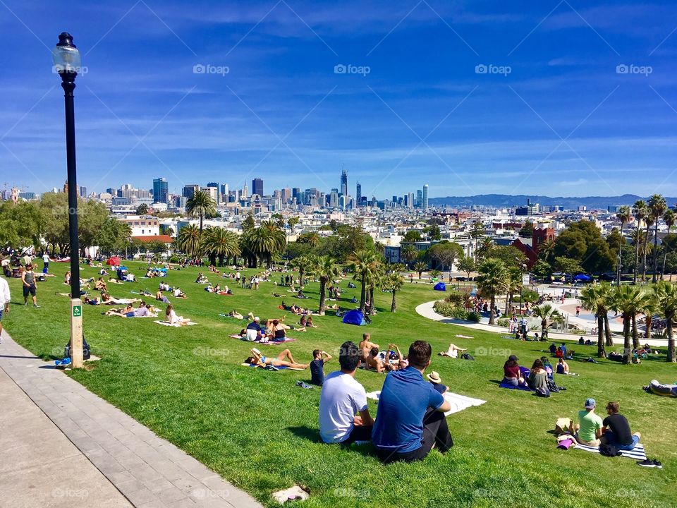 People lounge on the top of a green hill at the top of Mission Dolores Park in San Francisco. The San Francisco city skyline juts up in the distance. 