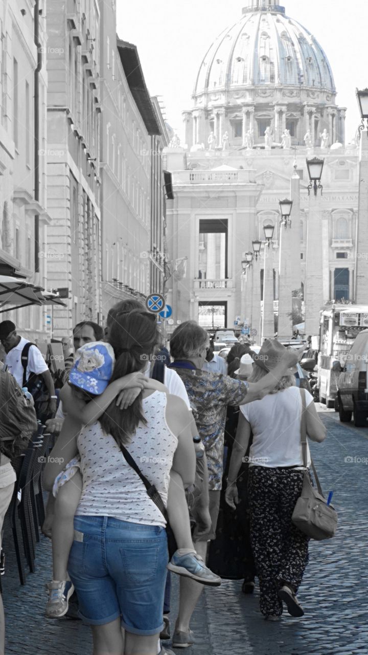 Mum walks, carrying her child in arms. In background: St. Peter's Basilica (Vatican).