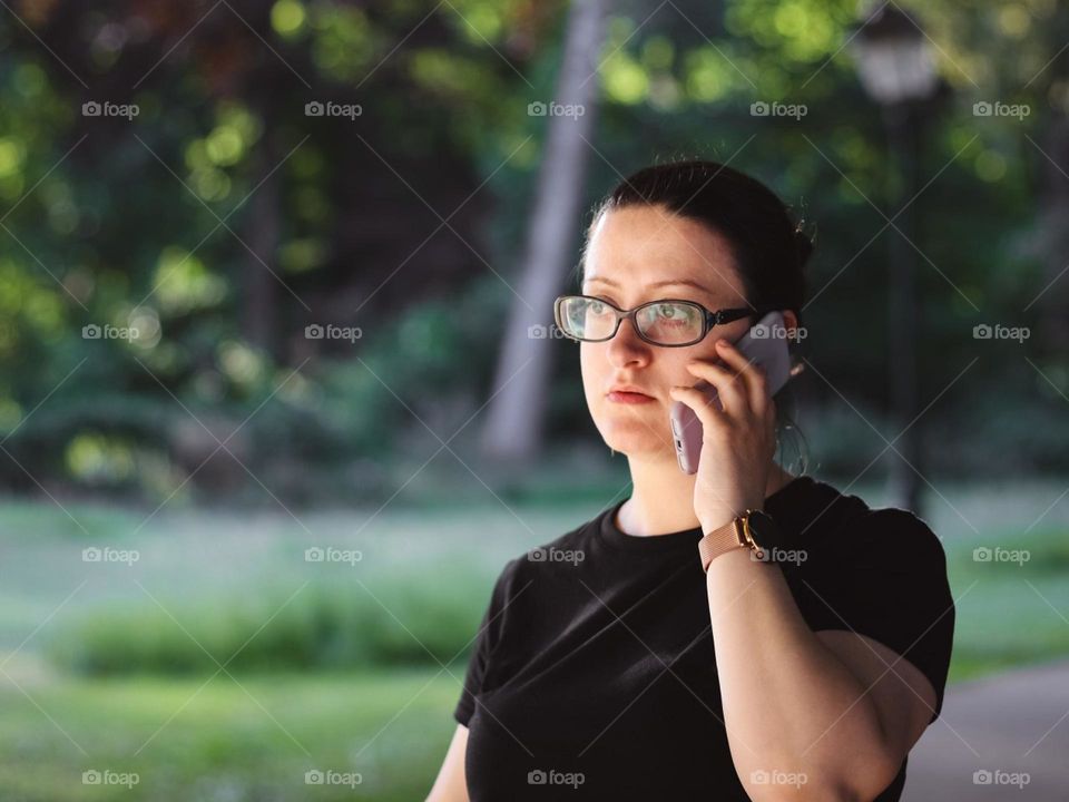 Portrait of one young beautiful middle-aged brunette Caucasian woman talking on a mobile phone and walking in the park on a sunny summer day, close-up side view.