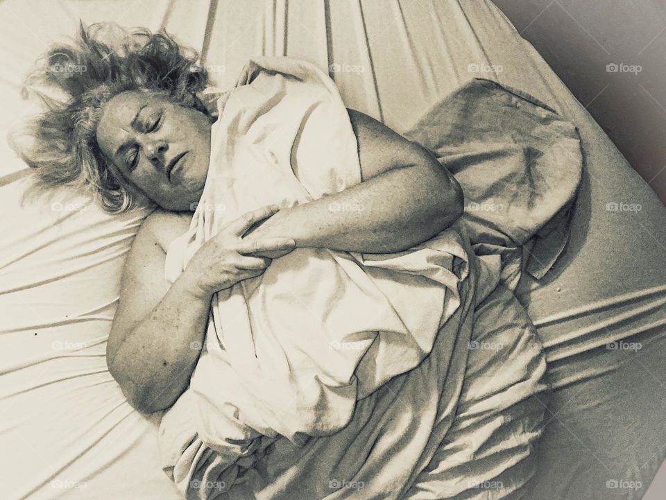 Silver tone self-portrait of a woman lying on a bed tightly bundled in a sheet. Her hair is messily splayed around her face, her eyes are closed and her hands are tightly clenched together. Her whole body appears tense and rigid.