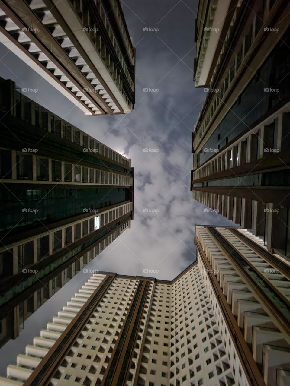 Framed by towering skyscrapers, the night sky becomes a canvas of ethereal clouds and moonlight. This upward perspective transforms the urban architecture into a geometric masterpiece.