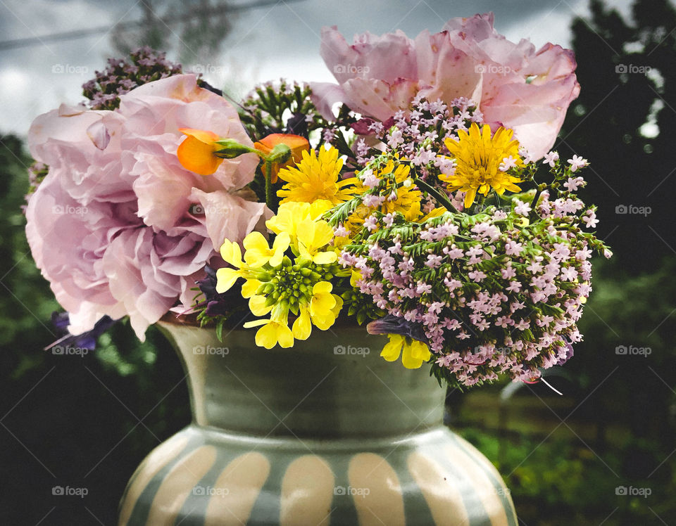 A green and cream vase containing springtime wildflowers sits on a balcony. The flowers work in colourful contrast to the rainy sky beyond