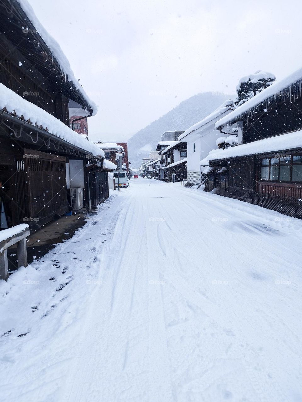 A peaceful snow-covered village street, with icicle-lined rooftops and a misty mountain in the background, capturing winter’s quiet beauty.