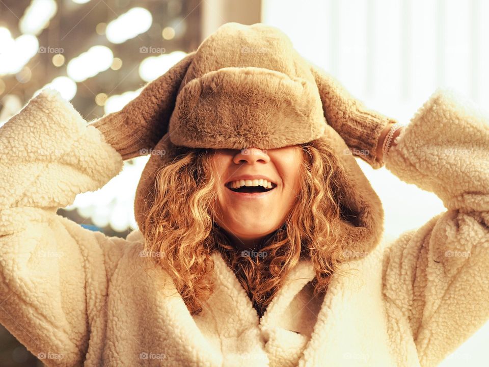 Young beautiful woman with curly hair in fur coat and hat smiling with lights on a background