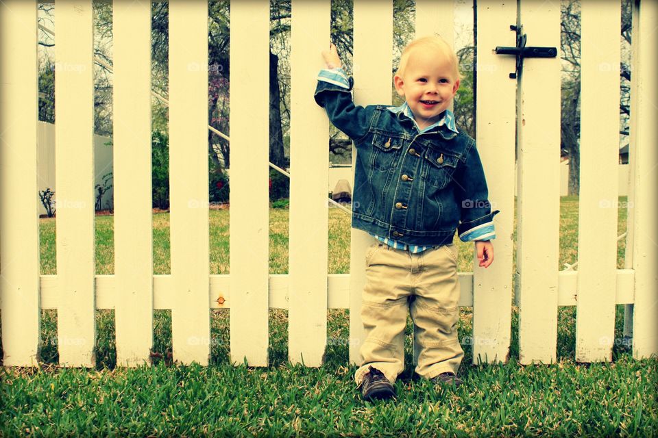Toddler boy standing on grass near wooden fence