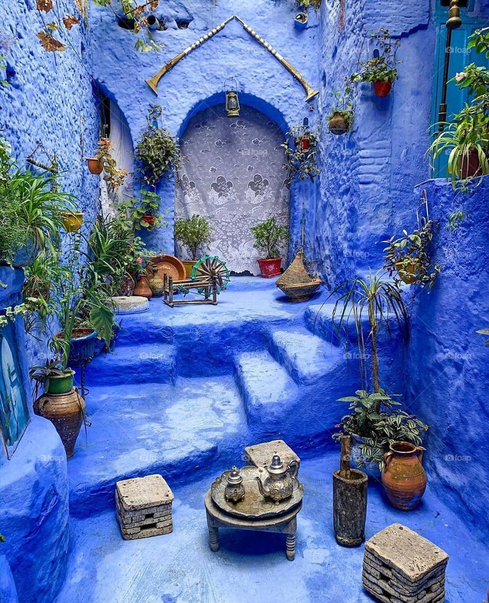 The image portrays a vibrant interior courtyard painted in striking shades of blue, featuring numerous traditional elements inspired by Moroccan decor. The walls, floor, and stairs are painted in captivating blue tones. Potted plants, both hanging an