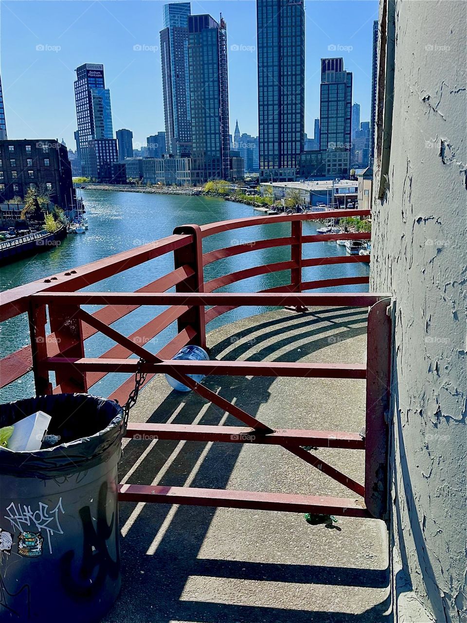 This is the center outpost of the “Pulaski Bridge” at “Newtown Creek” that connects “Greenpoint”, Bklyn to LIC. Across the “East River” we see “Manhattan”. The afternoon sun casts dramatic shadows onto the asphalt platform. 2024. Hypnotic Productions