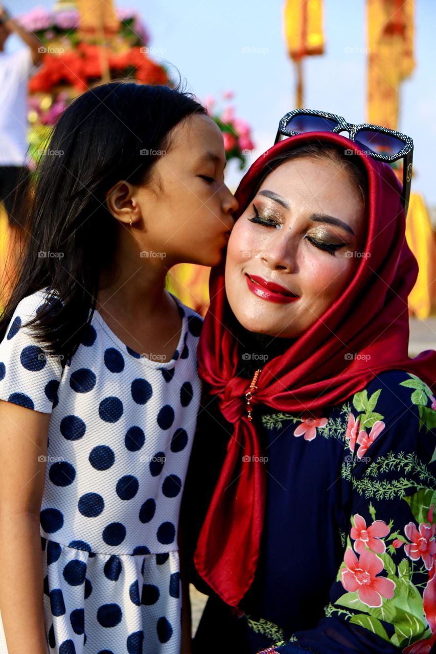 Portrait of a beautiful mother wearing a red hijab wearing a floral patterned robe and black glasses taking a photo with a happy expression being kissed by her daughter.