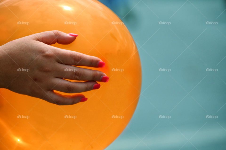 A hand with pink long pointed fingernails holds a bulging orange balloon