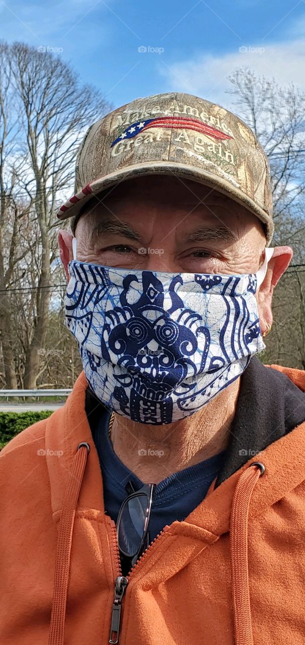 Man wearing a MAGA hat and face mask with what looks like a face on it. He's outside on a sunny chilly day.