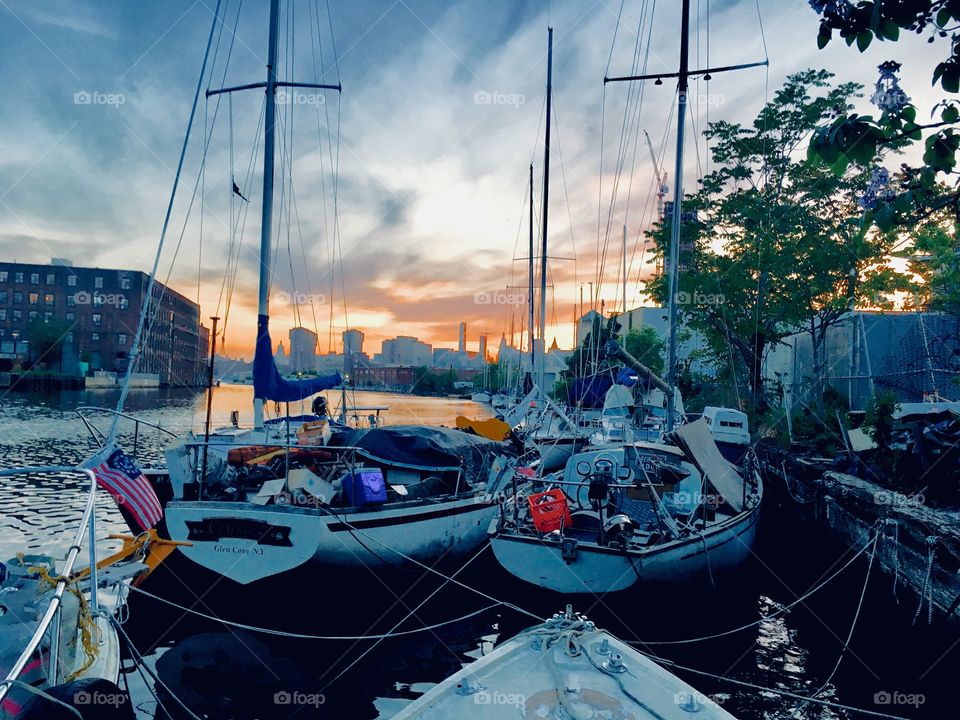 Sailboats in the East River at Newtown Creek, Long Island City, Queens, NY at twilight time. The cityscape at the horizon is the Manhattan skyline as well as parts of Queens. The sun’s last golden rays reflect in the clouds. 2018 Hypnotic Productions