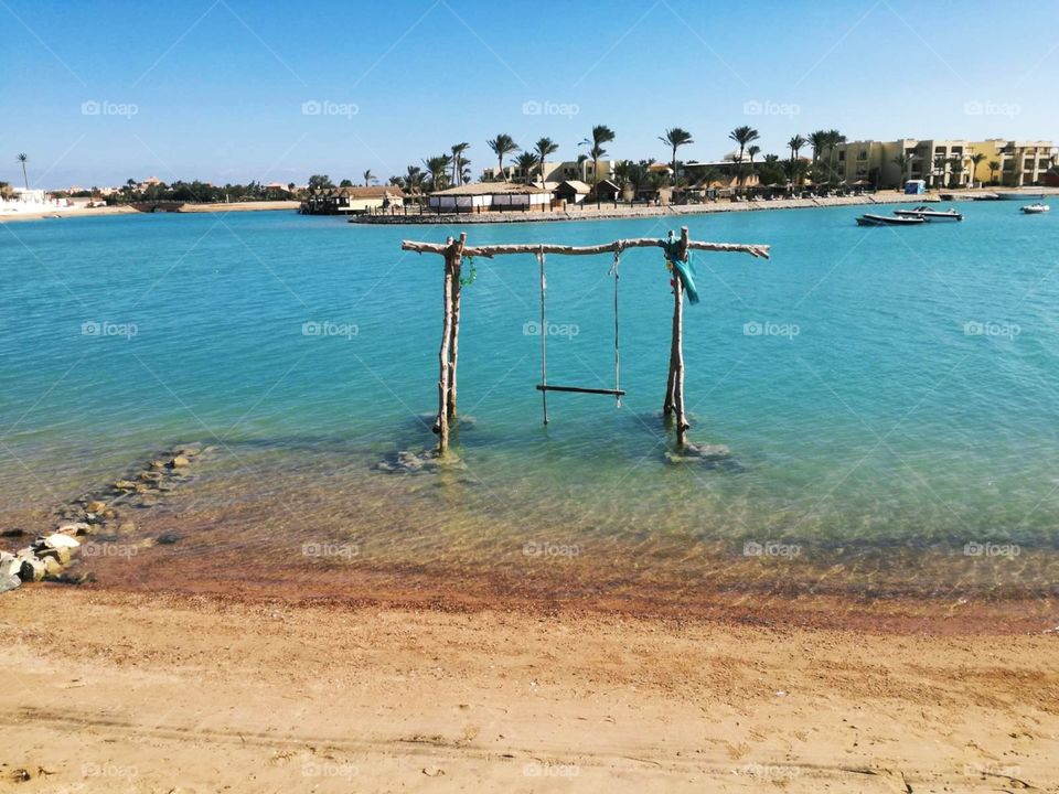 An interesting wooden swing in a beautiful blue sea. In amazing background there are boats, palms and houses.