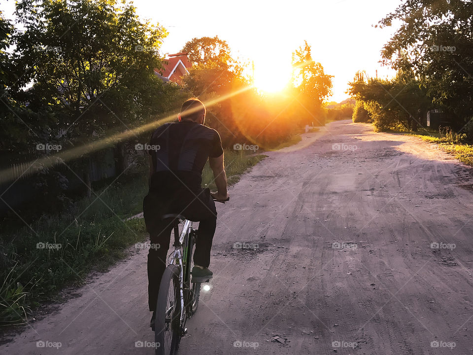 Young man riding a bicycle towards the sunset light by the village road 