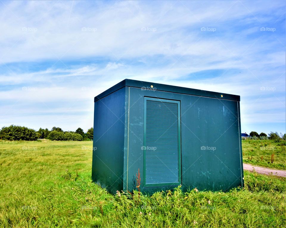 green rectangle electricity transformer house in an empty green field with blue sky and white clouds wide angle picture