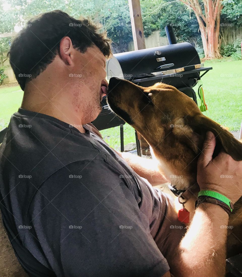 This man is getting some wet doggie kisses from his super cute hound dog while sitting outdoors. 