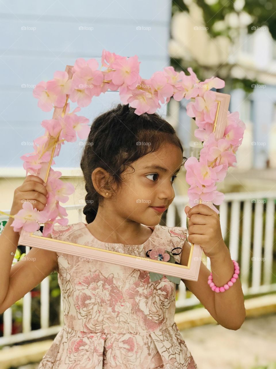 Beautiful baby girl wearing pink gown and pink bracelet to her hand holds a pink photo frame along with pink flowers and looking other side 