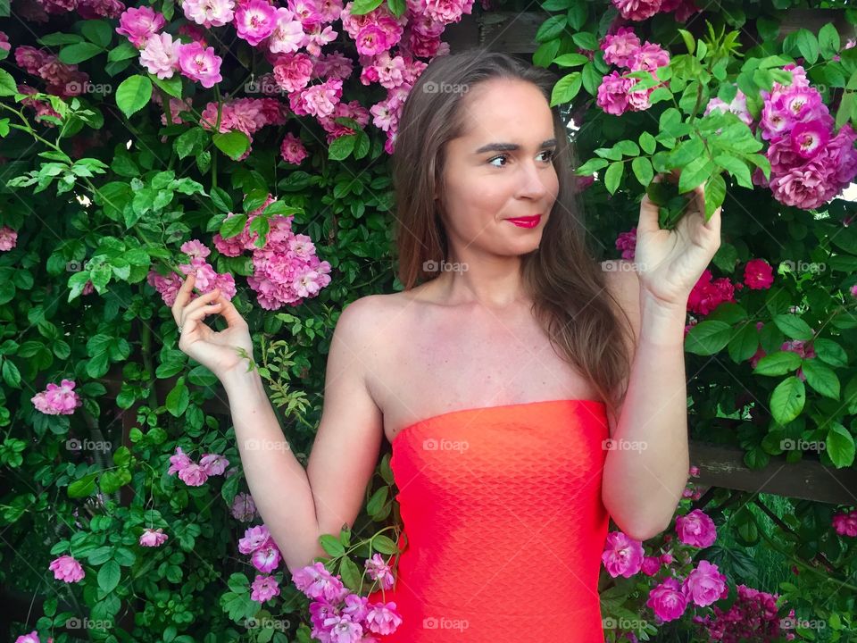 Portrait of pretty woman in red dress standing against wall of pink roses