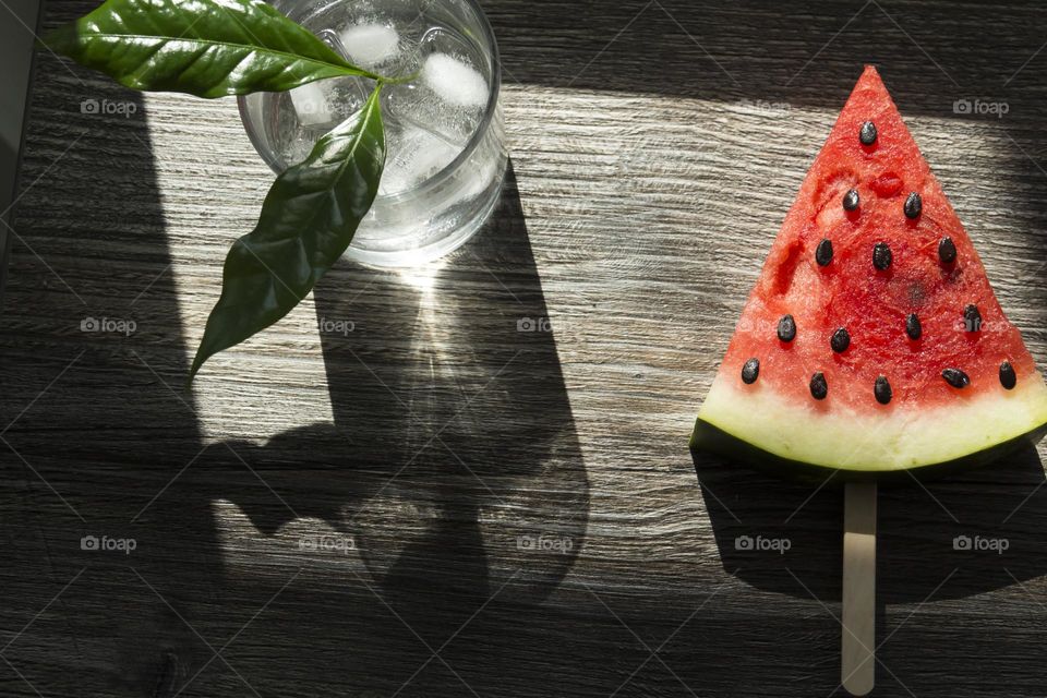 A piece of ripe, red watermelon and a cold cocktail with ice and green leaves are on a wooden table. Summer drinks and snacks.