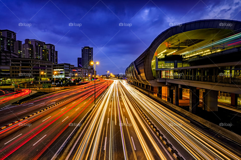 Light trail of busy traffic with LRT station railway, office building, and dark blue sky at the background. Location is in Puchong, Malaysia