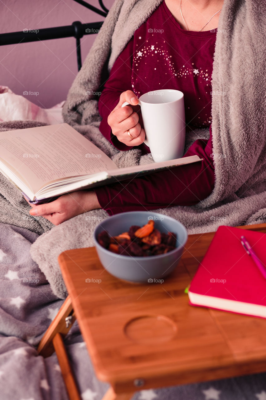 Woman enjoying the reading a book and drinking coffee at home. Young woman sitting in bed, wrapped in blanket, holding book, relaxing at home. Candid people, real moments, authentic situations